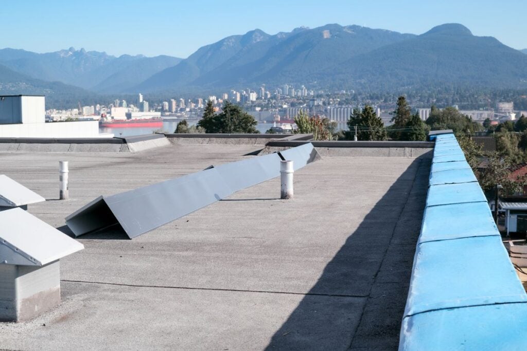cap sheet A flat rooftop with cap sheet material, metal vents, and a blue-edged barrier overlooks a city of tall buildings and forested mountains in the background, all beneath a clear sky—an urban scene that captures modern flat roofs in 2026.