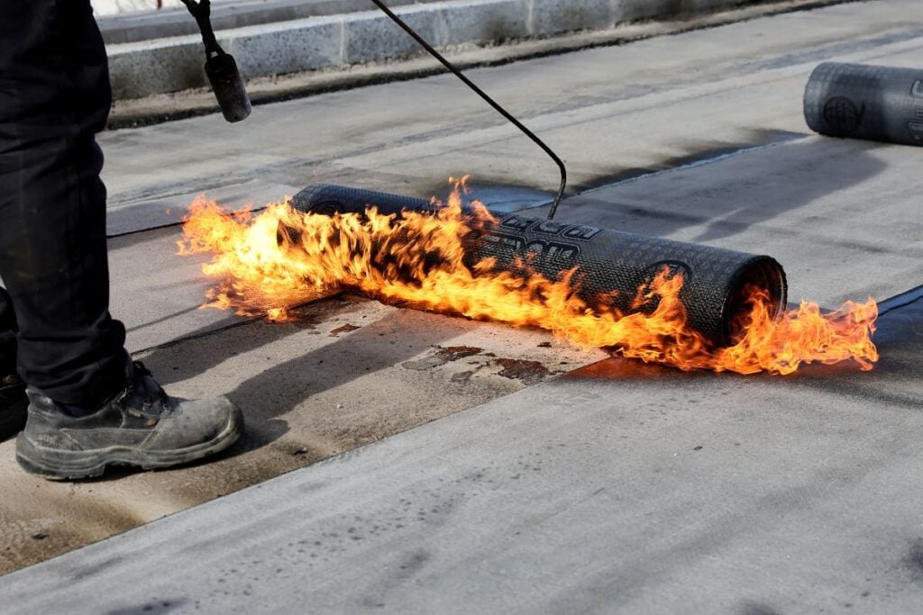cap sheet A person uses a torch to apply a cap sheet, with visible flames heating the material as it is unrolled across a flat roof surface—a common method for flat roofs expected to endure through 2026.
