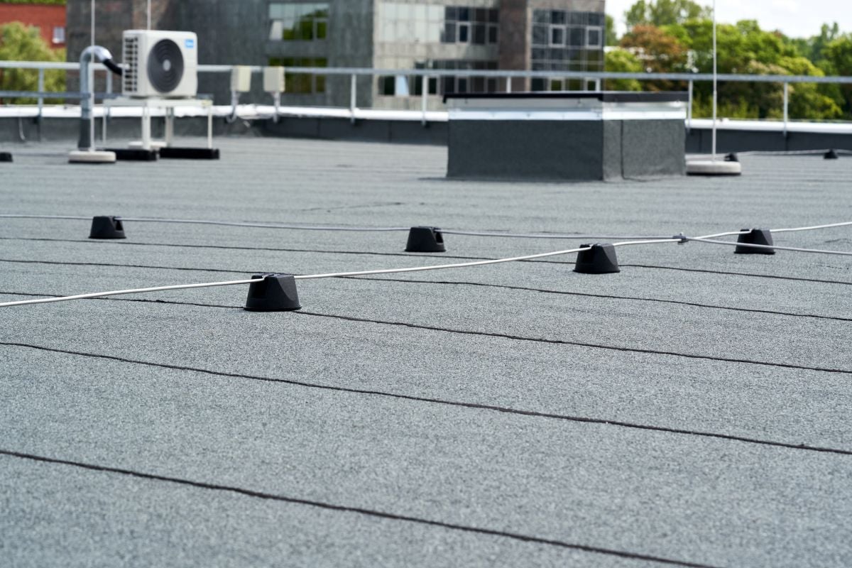 cap sheet A flat commercial rooftop with a cap sheet surface, safety cable system mounted on small black stanchions, and HVAC equipment and buildings in the background—an ideal example of modern flat roofs for 2026.