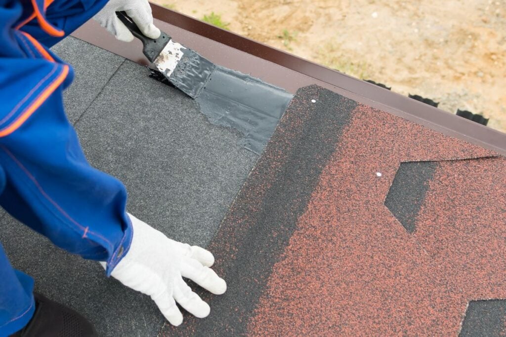 A person wearing white gloves and a blue uniform applies black roofing tar with a brush to seal shingles at the edge of a rooftop as part of regular roof maintenance.