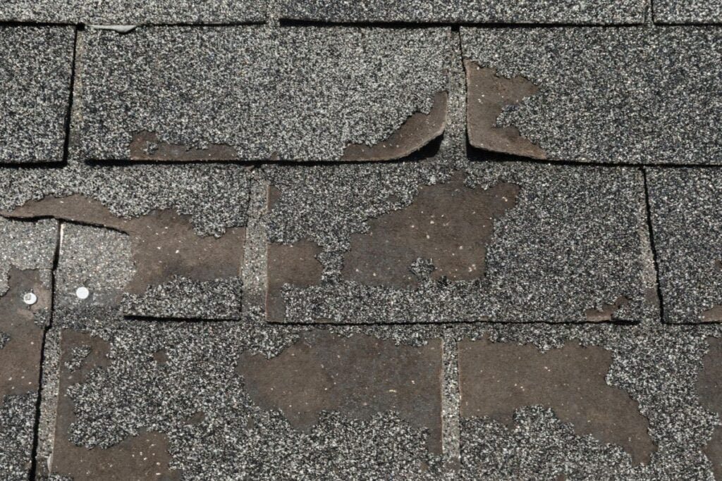 asphalt roof repair Worn and damaged asphalt roof shingles with patches of missing granules, exposing the underlying material and some bent shingle edges. Visible roof issues include two exposed nails on the left side, signaling a need for prompt asphalt roof repair.