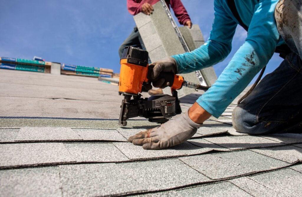 asphalt roof maintenance A worker wearing gloves uses a nail gun to install asphalt shingles on a roof as part of asphalt roof maintenance, while another person in the background carries more shingles under a clear blue sky.