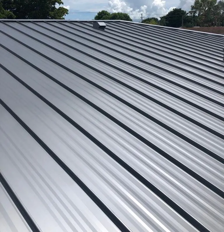 A close-up view of a shiny, silver metal roofing Cornelius NC with raised seams, photographed during the day with a partly cloudy sky and trees visible in the background.