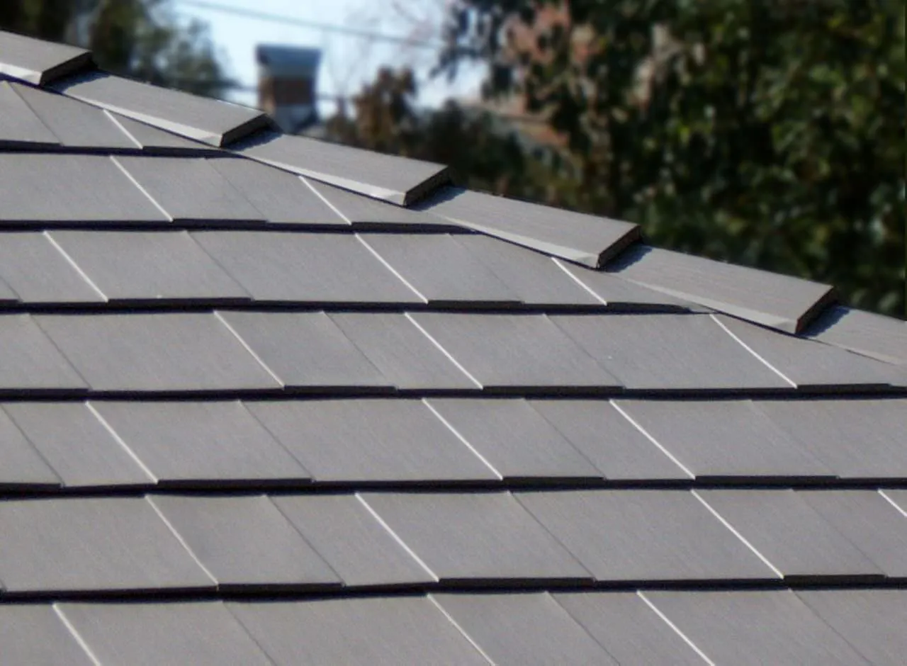 Close-up of a roof with dark gray, rectangular solar shingles arranged in neat rows; metal roofing Cornelius NC solutions blend seamlessly, while trees and sky are blurred in the background.