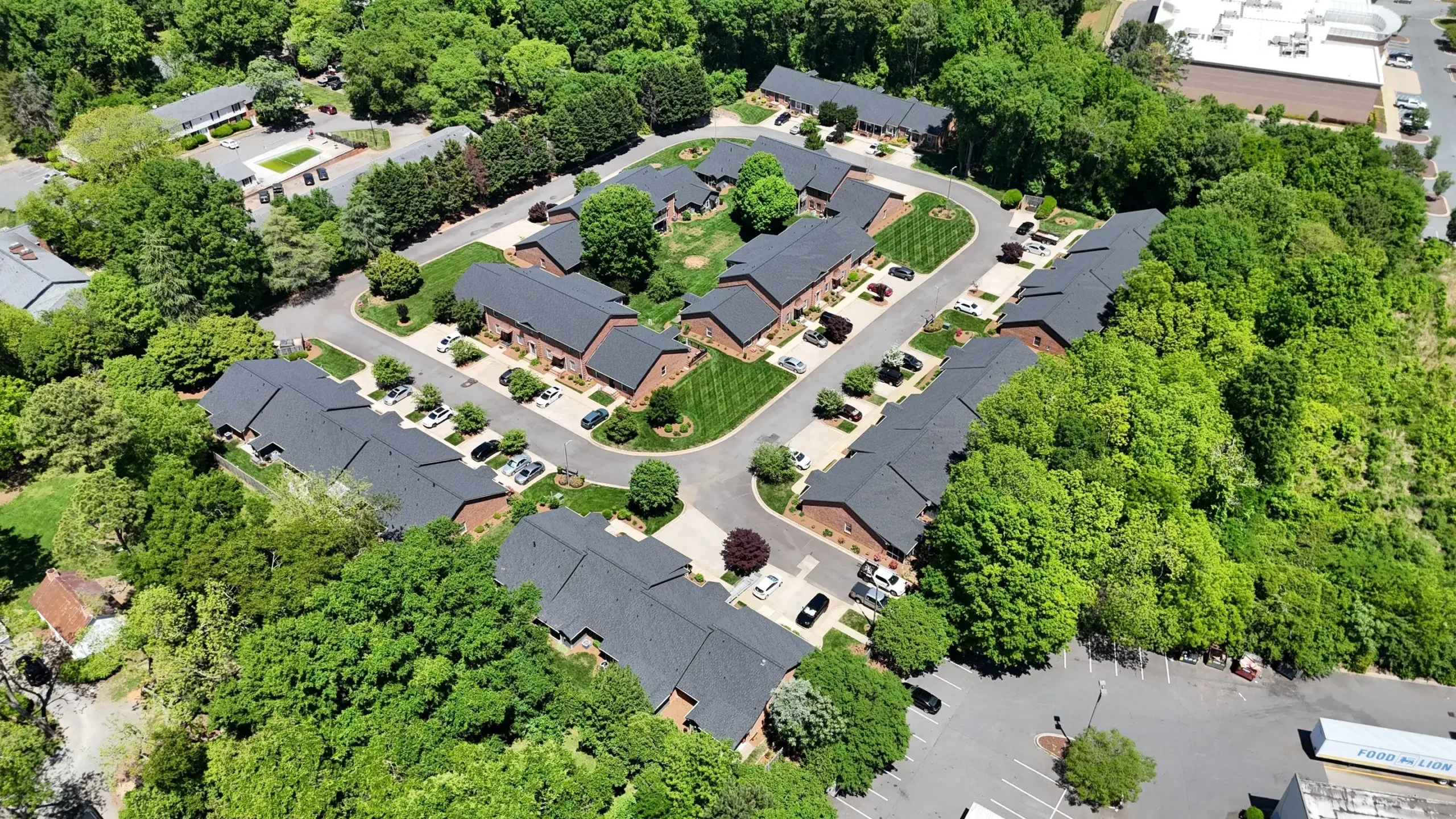 Aerial view of a residential complex surrounded by dense trees, with multiple dark-roofed buildings recently undergoing SouthPark Charlotte roof replacement, parking spaces, and a circular road layout. Nearby, commercial buildings and parking lots are partially visible.