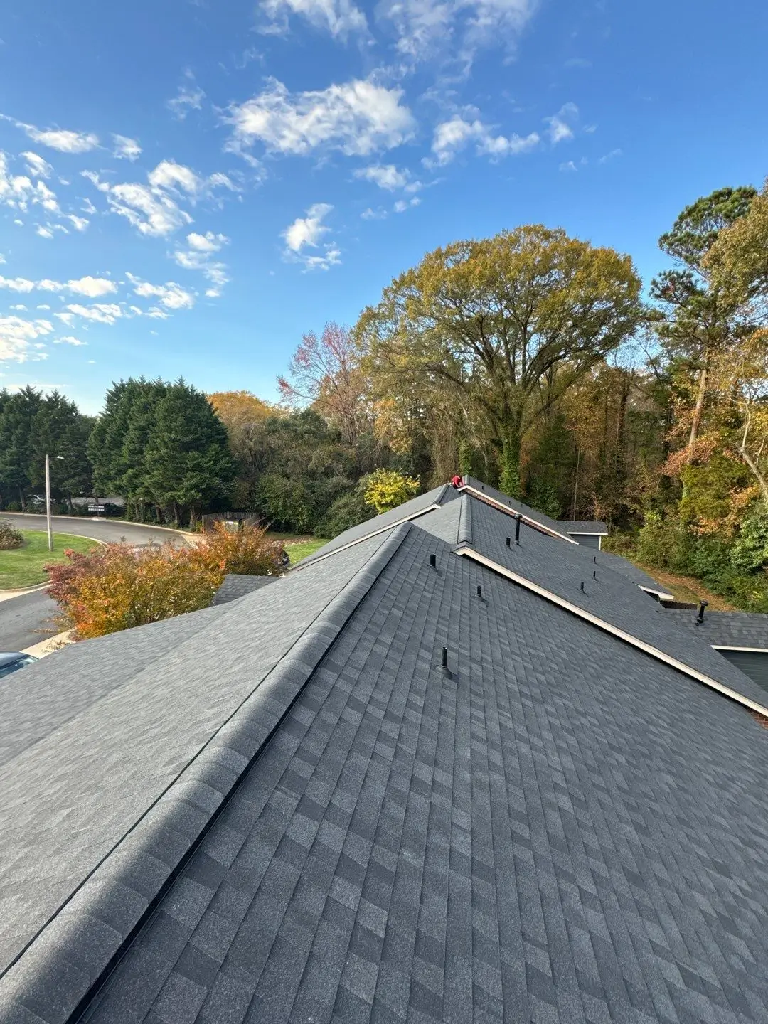 View from a rooftop with new dark shingles after a recent SouthPark Charlotte roof replacement, looking over a suburban area with trees, a winding road, and a partly cloudy blue sky.