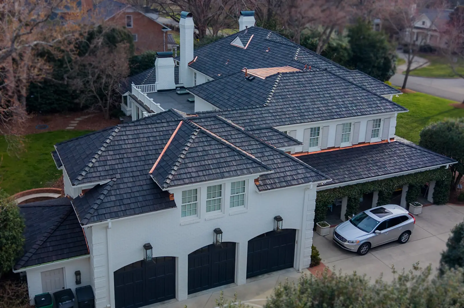 A large two-story house with dark roof tiles, light exterior walls, black garage doors, and a silver car parked in the driveway is surrounded by trees and greenery, showcasing the elegance of a recent luxury roof replacement in Greensboro, NC.