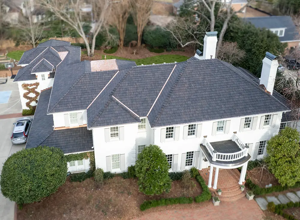 Aerial view of a large two-story white house with dark gray shingles, white columns, a balcony above the entrance, and surrounding greenery and trees—showcasing the elegance and quality you can expect from a luxury roof replacement in Greensboro NC.