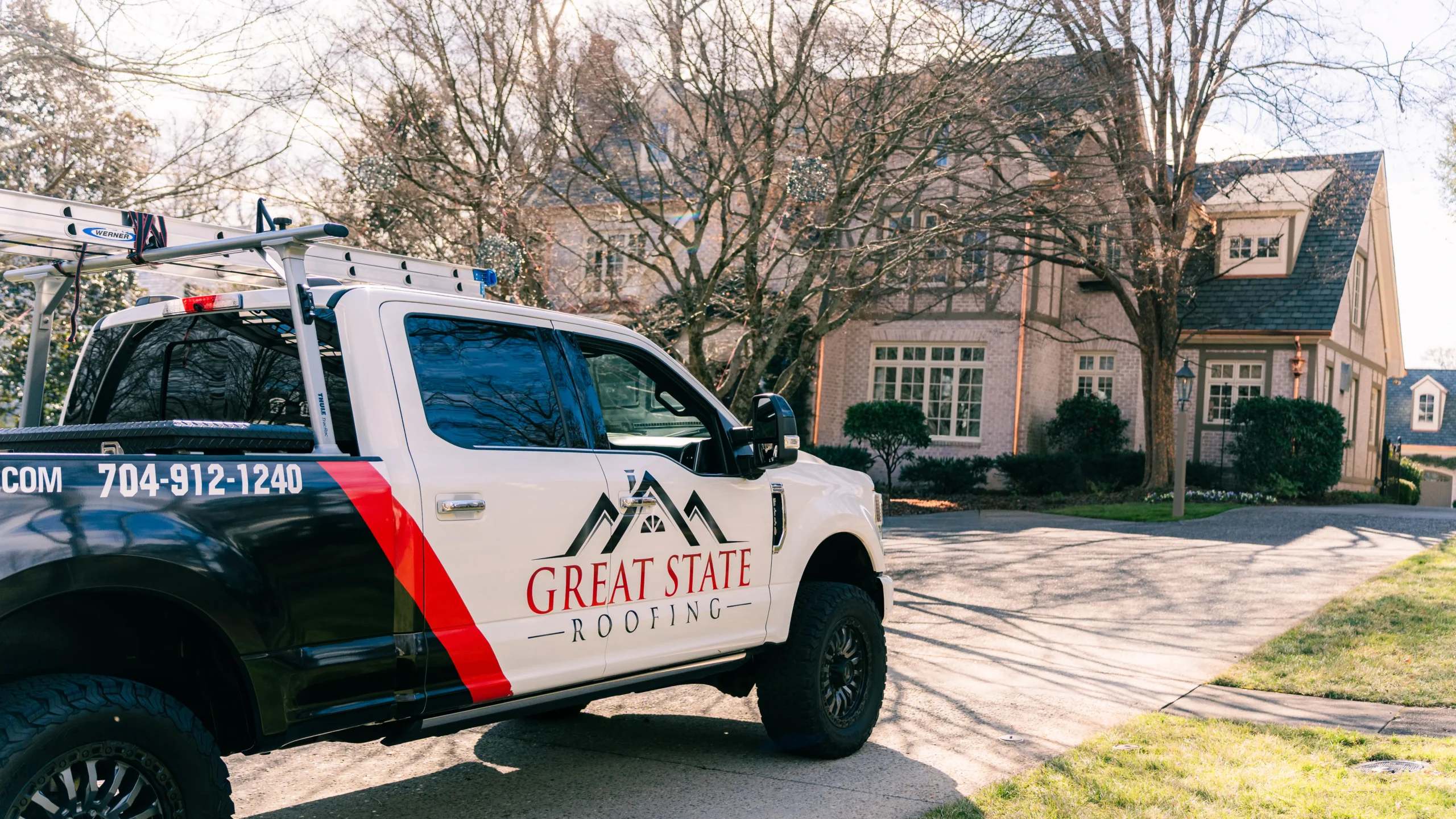 A Great State Roofing truck is parked in front of a large suburban brick house with trees and bushes in the yard on a sunny day, showcasing the trusted expertise of roofing Greensboro NC homeowners rely on.