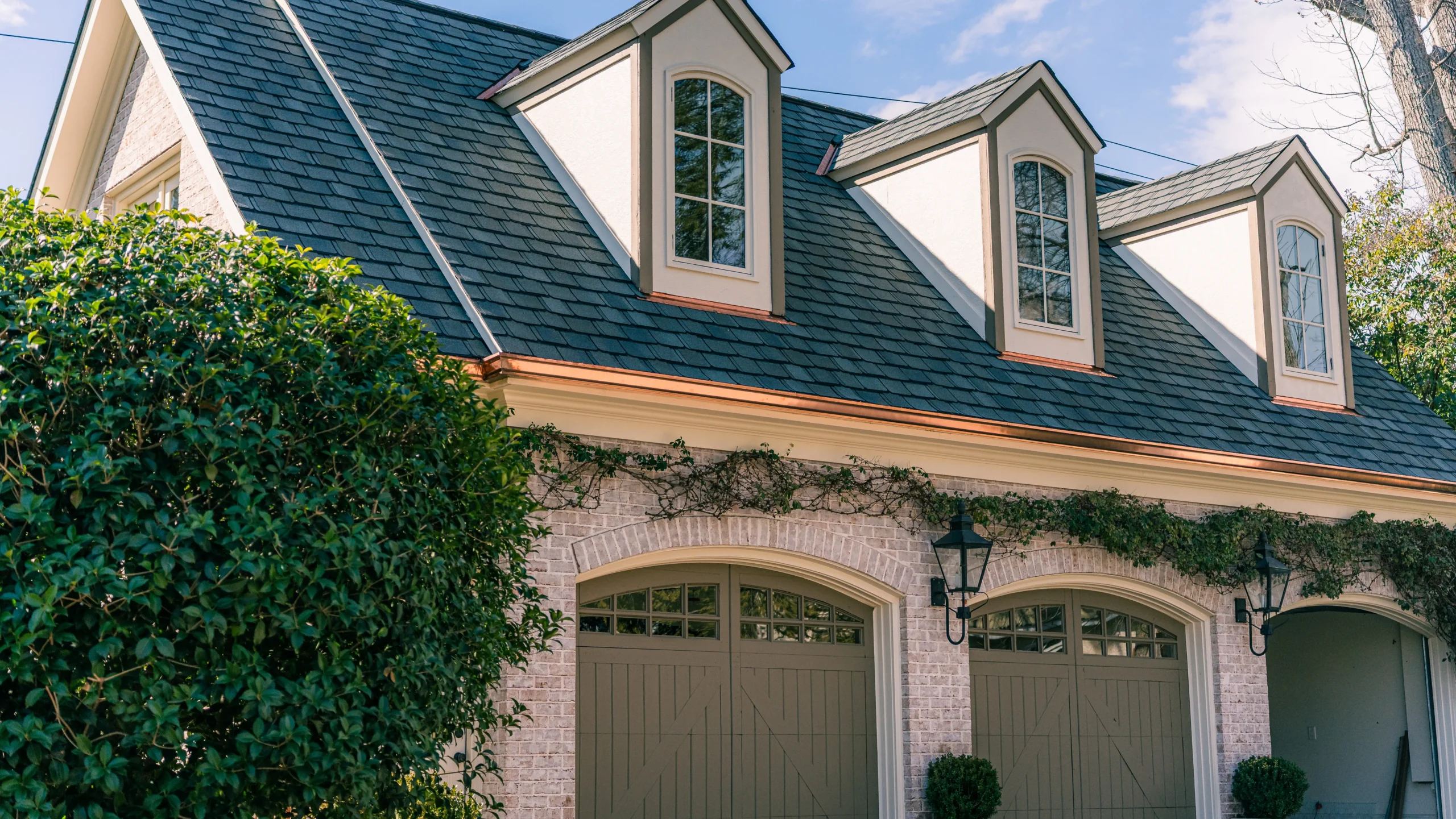 A two-car garage with light gray doors, brick walls, black shingles installed by experts in roofing Greensboro NC, three dormer windows, and green ivy growing above the doors. Bushes and trees surround the building under a clear sky.