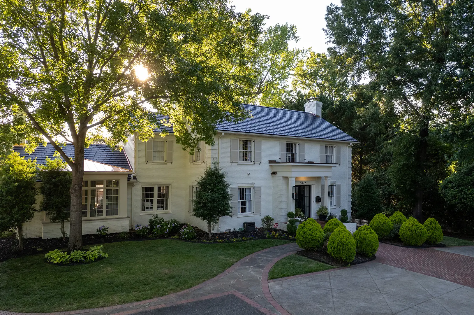 Large two-story white brick house with a dark roof, surrounded by trees and shrubs. A curved driveway leads to the front entrance, and sunlight filters through the leaves, highlighting the impeccable results of a recent luxury roof replacement in Greensboro, NC.