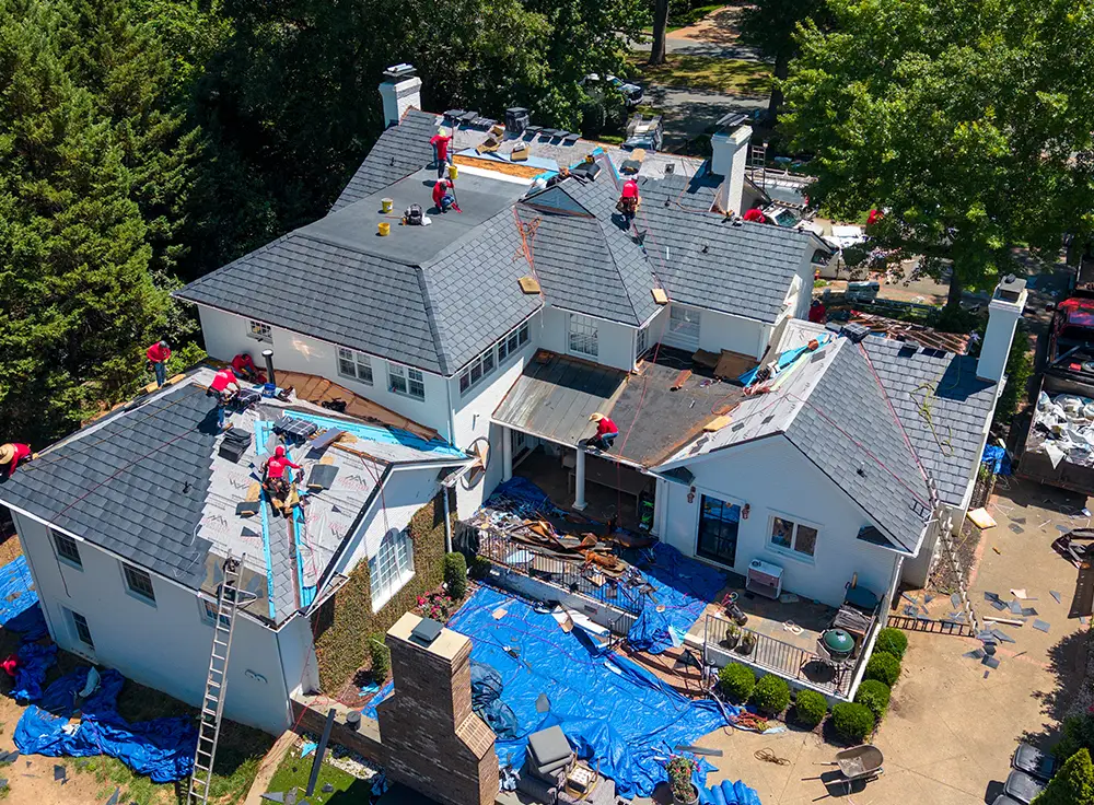 Aerial view of roofers in red shirts expertly repairing and replacing shingles on a large house roof, with tools, ladders, and blue tarps covering parts of the ground and roof areas—demonstrating a meticulous luxury roof replacement in Greensboro, NC.