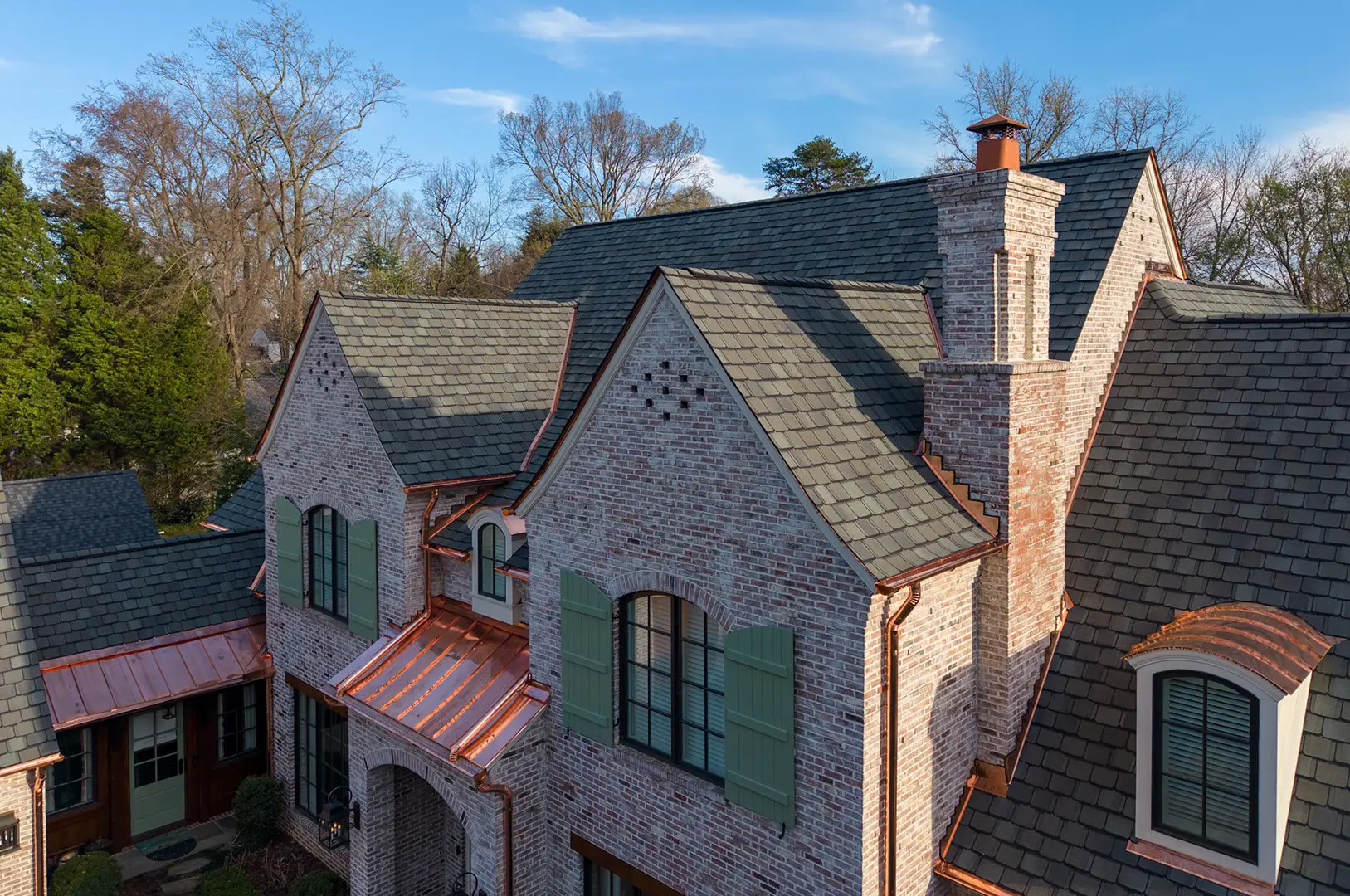 Brick house with green shutters, gray shingle roof, copper roofing accents, and large chimneys. Trees without leaves surround the home under a blue sky with some clouds, reflecting the timeless charm of Greensboro luxury roof replacement.