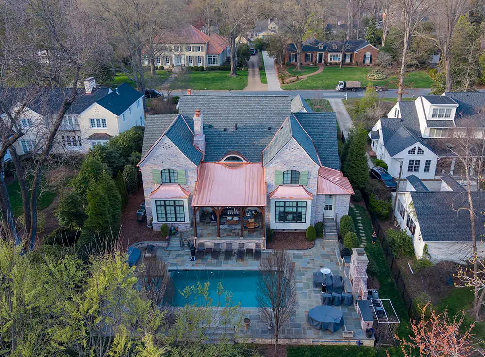 Aerial view of a large stone house with a pool in the backyard, featuring a Greensboro Luxury Roof Replacement, surrounded by trees and neighboring homes in a suburban area.