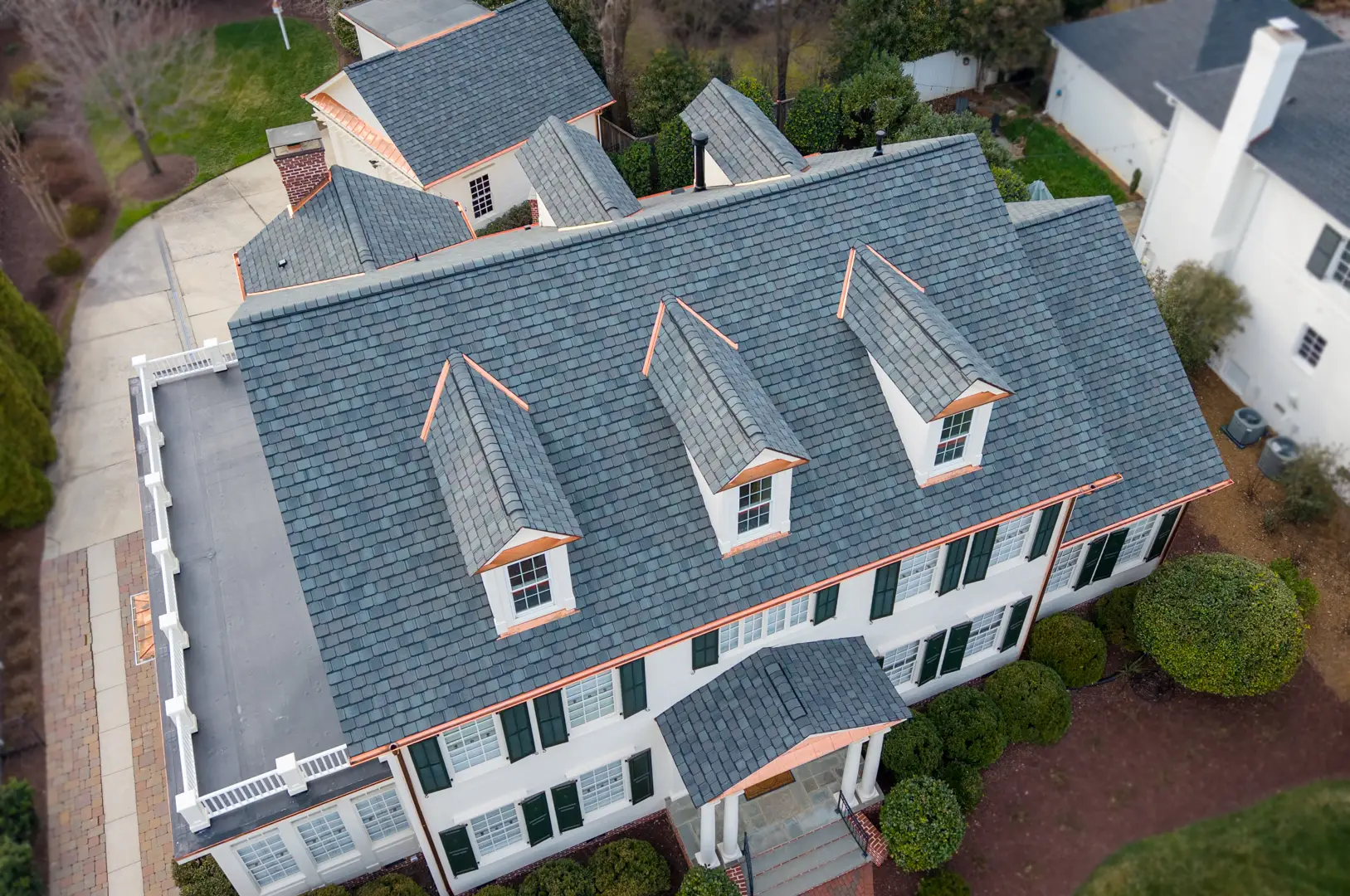 Aerial view of a large, two-story house with gray shingle roof, three dormer windows, white exterior walls, and green shutters, surrounded by bushes and trees—showcasing the impeccable results of a luxury roof replacement in Greensboro NC.