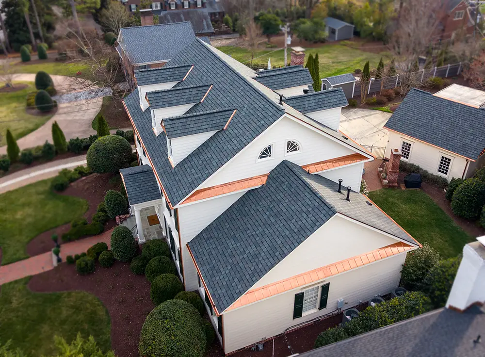 Aerial view of a large, light-colored house with a dark gray roof, dormer windows, and copper roof accents—showcasing the impeccable craftsmanship of a luxury roof replacement in Greensboro NC—surrounded by shrubs, trees, and paved walkways.