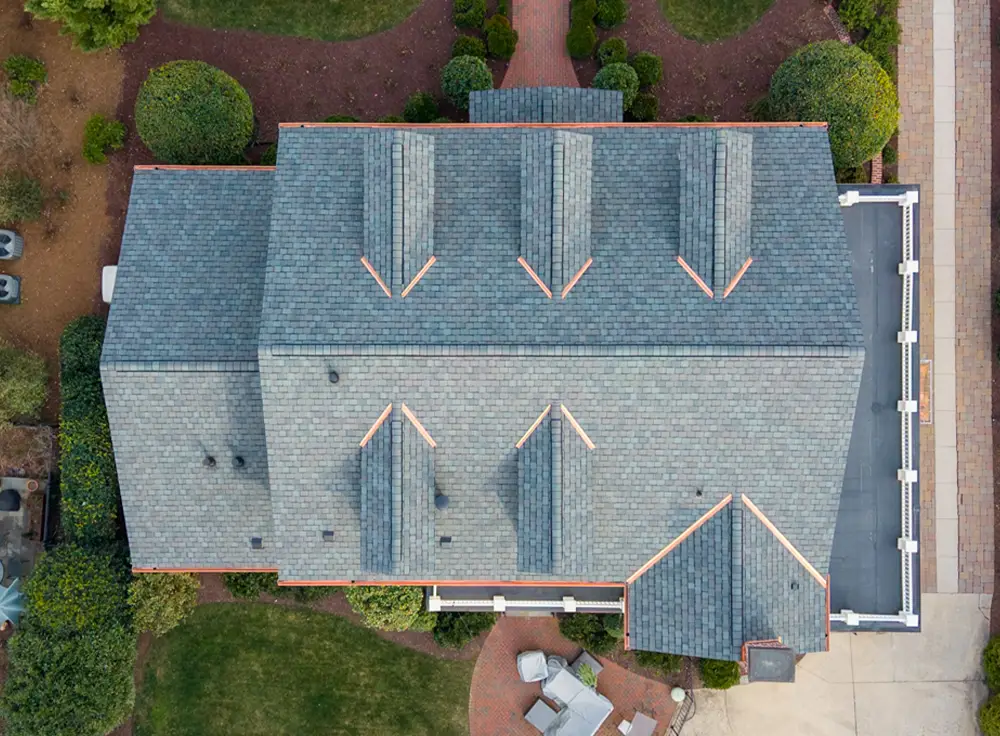 Aerial view of a gray-roofed house with multiple dormer windows, surrounded by green bushes, a walkway, and outdoor furniture on a patio—an impressive example of luxury roof replacement in Greensboro NC.