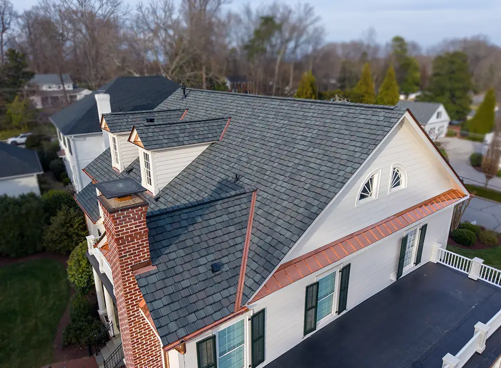 Aerial view of a house with gray shingle roofing, white siding, brick chimney, green shutters, and copper flashing—showcasing the attention to detail characteristic of a luxury roof replacement in Greensboro, NC. Surrounding area includes trees and neighboring homes.
