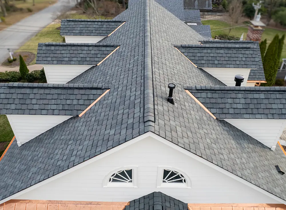 Aerial view of a large house roof with dark gray shingles, several dormer windows, copper flashing, and two black roof vents. The symmetrical design and sharply peaked ridge showcase the refined results of a luxury roof replacement Greensboro NC homeowners can trust.