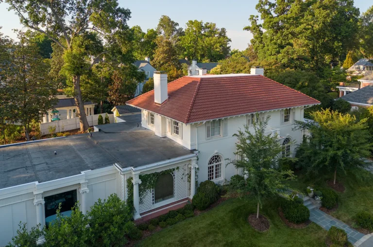 A large white two-story house with a Spanish tile roof replacement in Greensboro, NC features arched windows and is surrounded by trees on a sunny day. A flat-roofed extension is attached to the side, while pathways and trimmed bushes line the front yard.