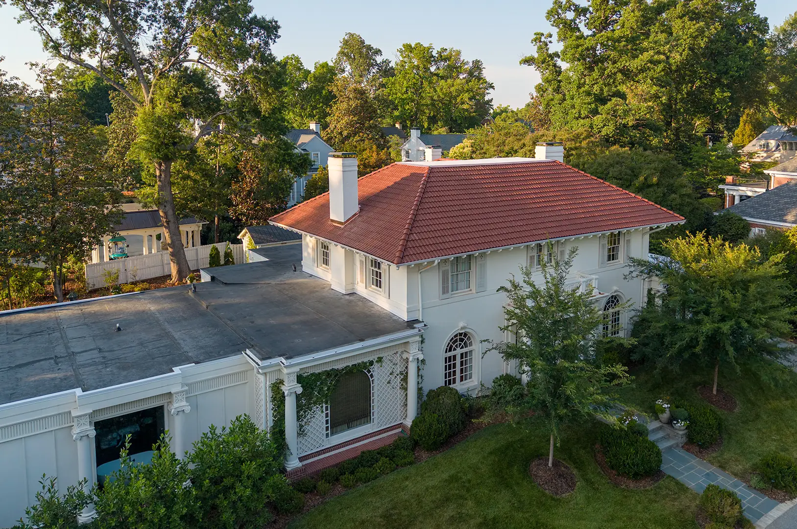 A large white two-story house with a Spanish tile roof replacement in Greensboro, NC features arched windows and is surrounded by trees on a sunny day. A flat-roofed extension is attached to the side, while pathways and trimmed bushes line the front yard.