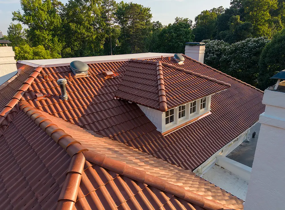 A rooftop view of a house with reddish-brown clay tiles, skylights, a dormer window, and surrounded by green trees under a clear sky—perfect inspiration for anyone considering Spanish tile roof replacement in Greensboro NC.