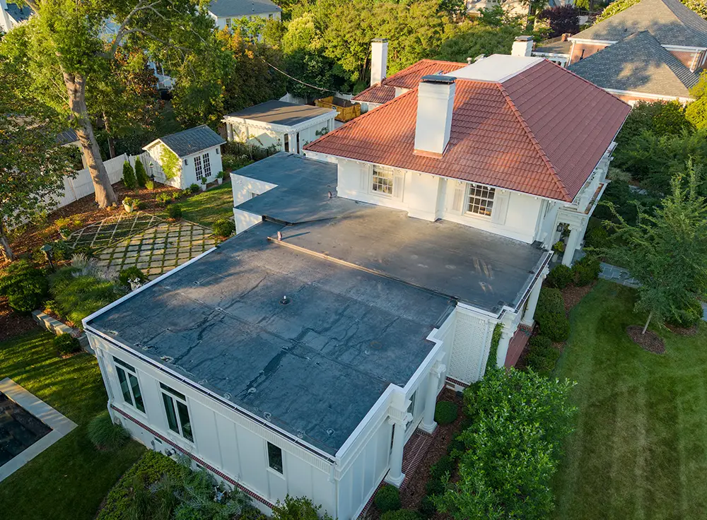 Aerial view of a large house with a red-tiled roof and an attached flat-roofed section, showcasing recent Spanish tile roof replacement in Greensboro, NC, surrounded by green lawns, trees, and garden structures.