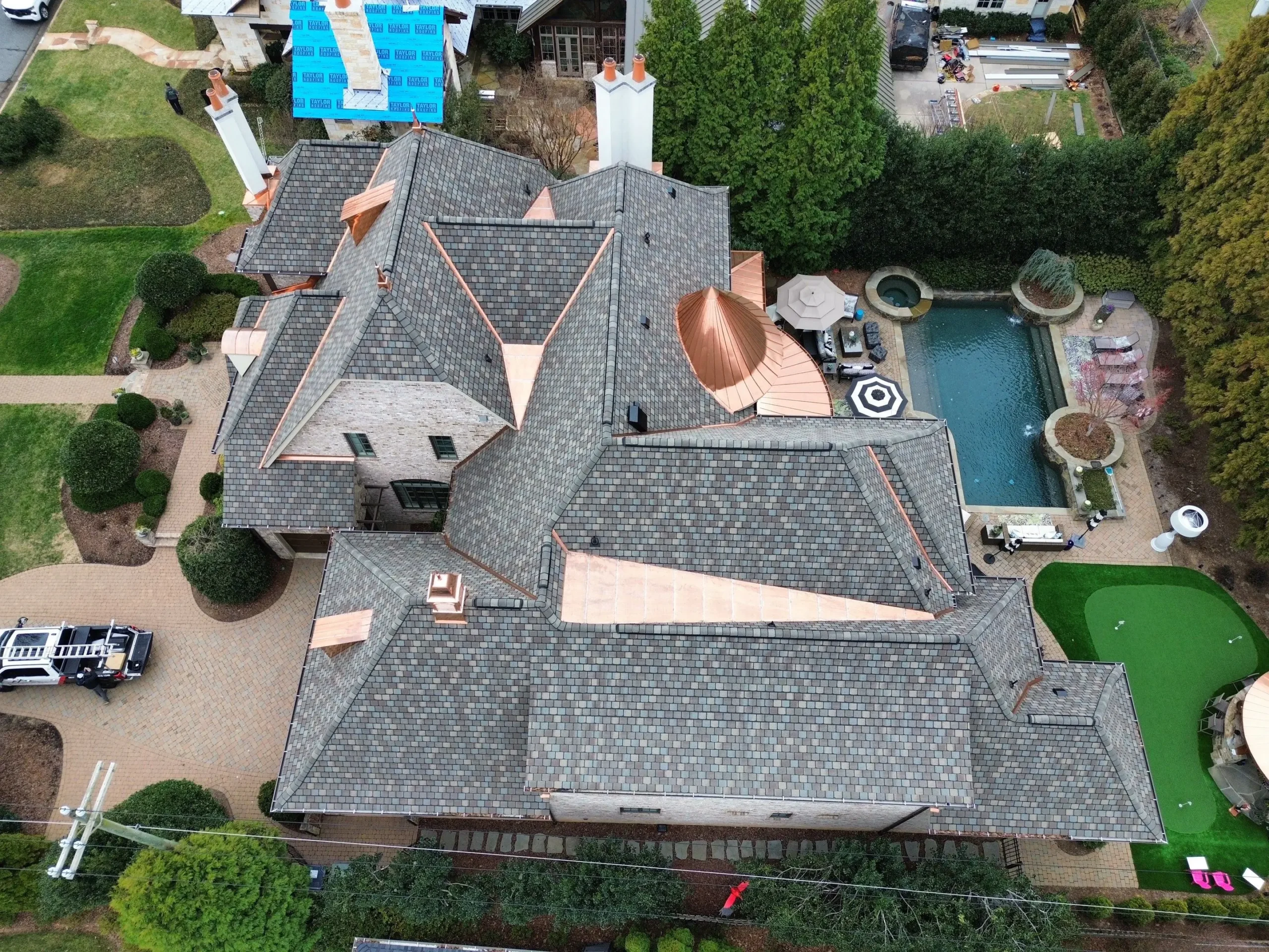 Aerial view of a large house with a complex roof, a copper dome, swimming pool, patio furniture, mini-golf area, and landscaping with bushes and trees. A blue construction tarp is visible on part of the roof, indicating ongoing roofing Greensboro NC work.