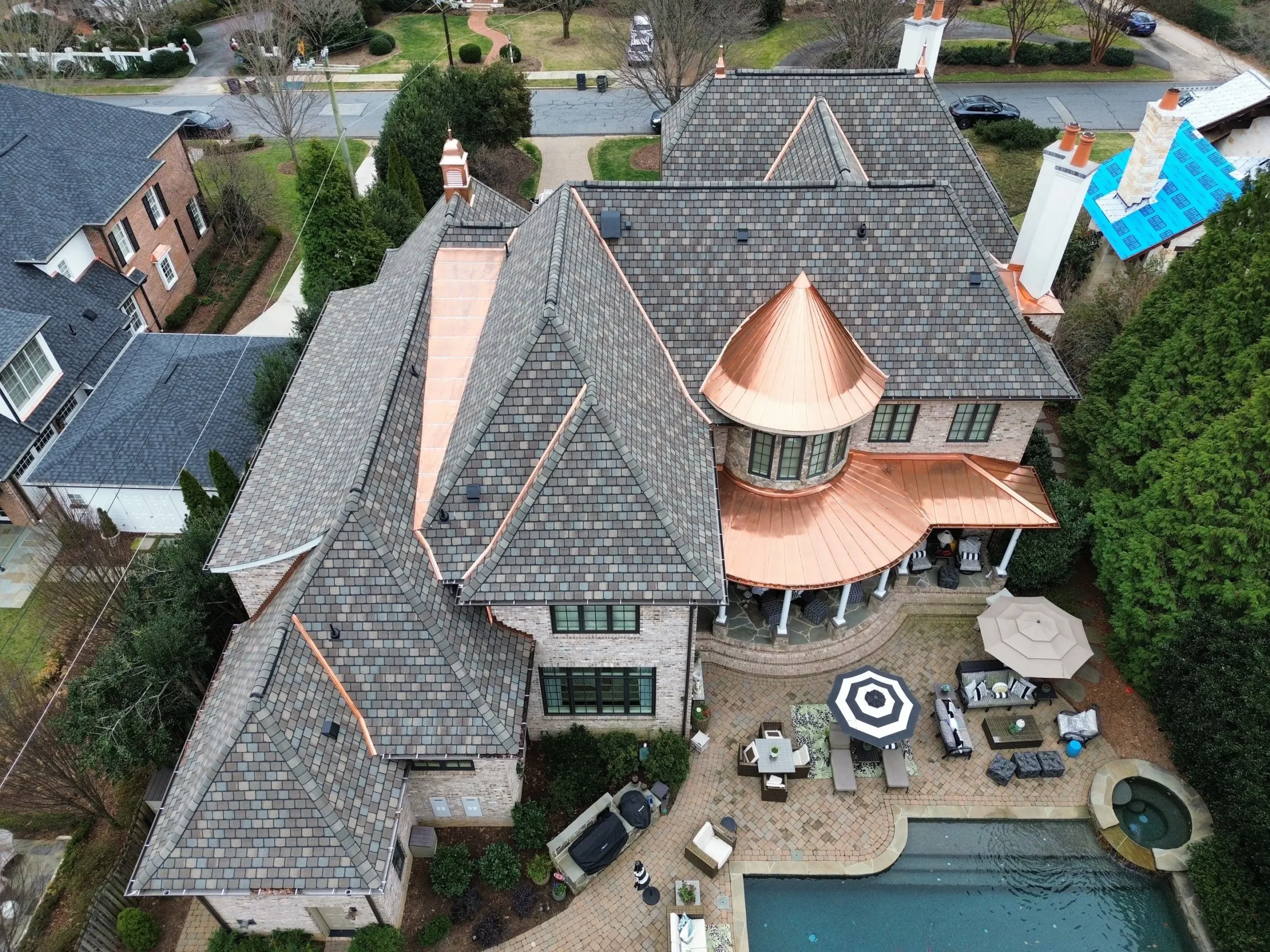 Aerial view of a large house with gray shingles and copper roofing Greensboro NC, featuring a backyard patio, seating areas, and a swimming pool with a hot tub. Trees and neighboring homes surround the property.