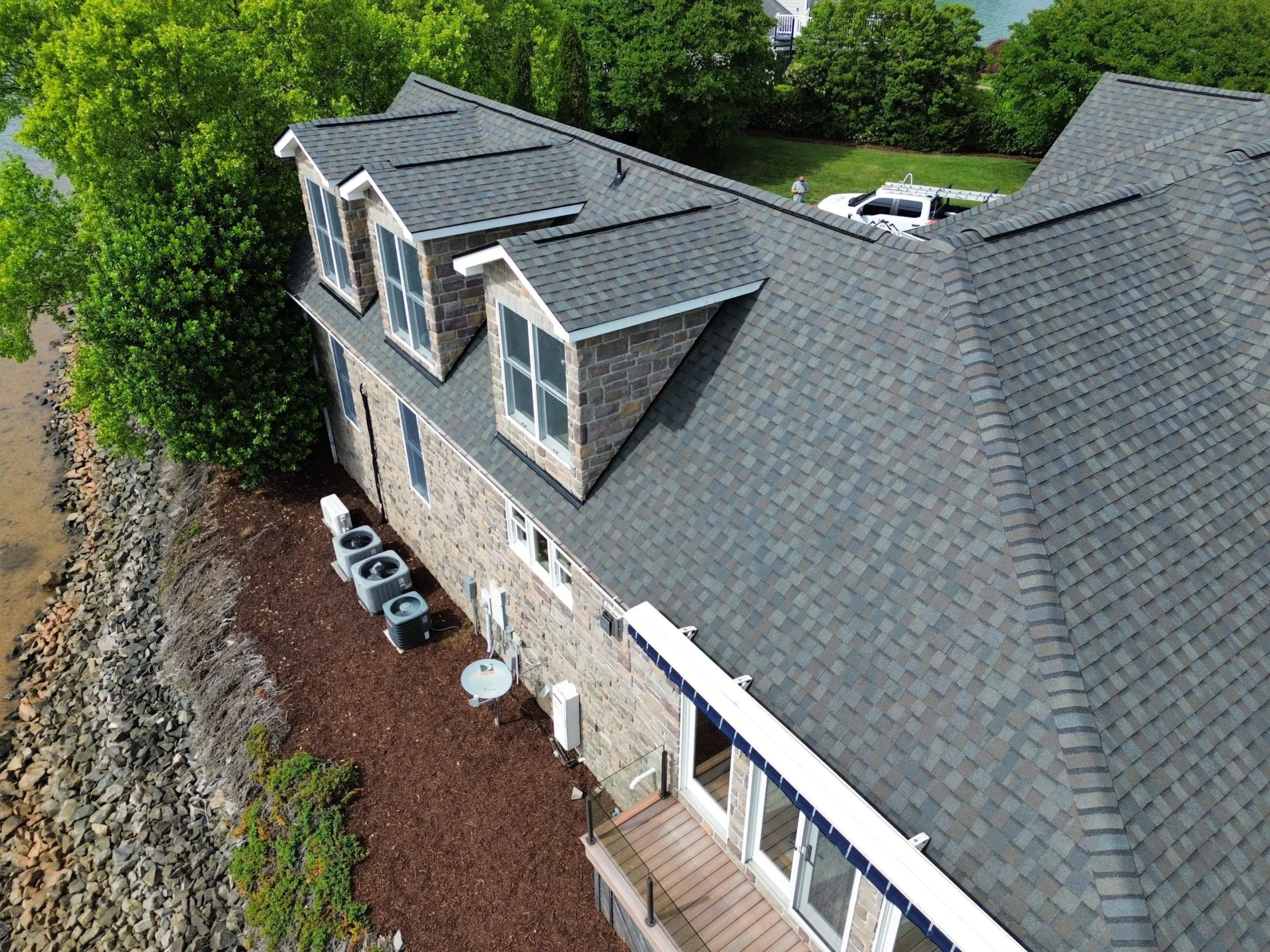 Aerial view of a gray-shingled, Brava Cedar Shake stone-sided house by water, with three dormer windows, air conditioning units beside it, and a wooden deck below surrounded by mulch and trees.