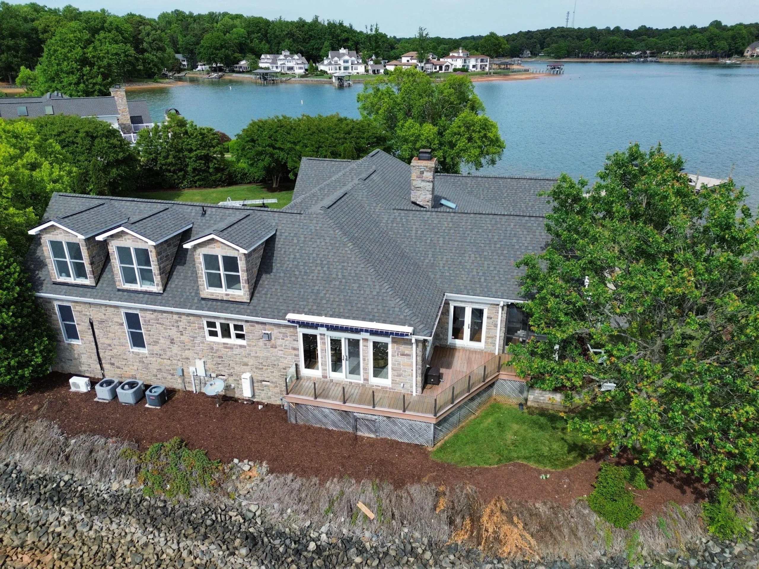 A large stone house with multiple dormer windows sits on a lakefront, its roof adorned with Brava Cedar Shake, surrounded by greenery and trees, with a wooden deck overlooking the water and nearby houses across the lake.