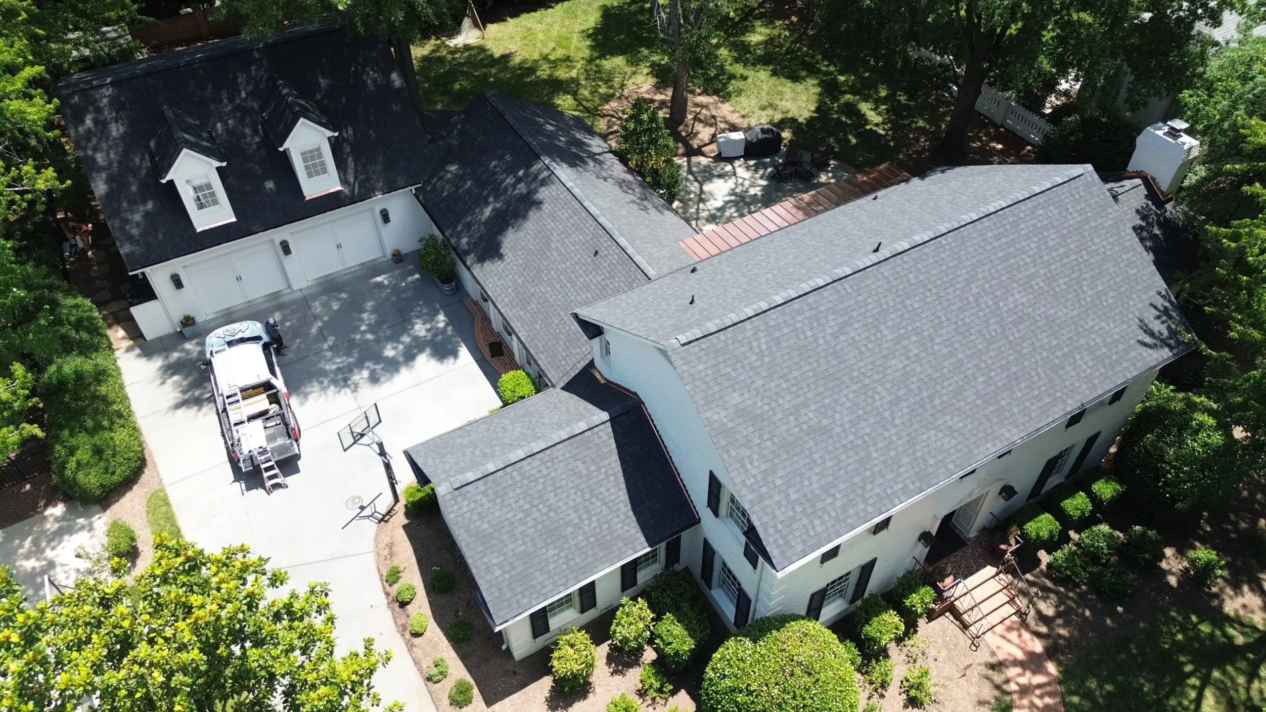 Aerial view of a large white house with a dark roof, attached garage, basketball hoop, boat on a trailer in the driveway, and landscaped yard with bushes and trees surrounding the property—showcasing the results of a recent SouthPark Charlotte roof replacement that enhances both durability and curb appeal.