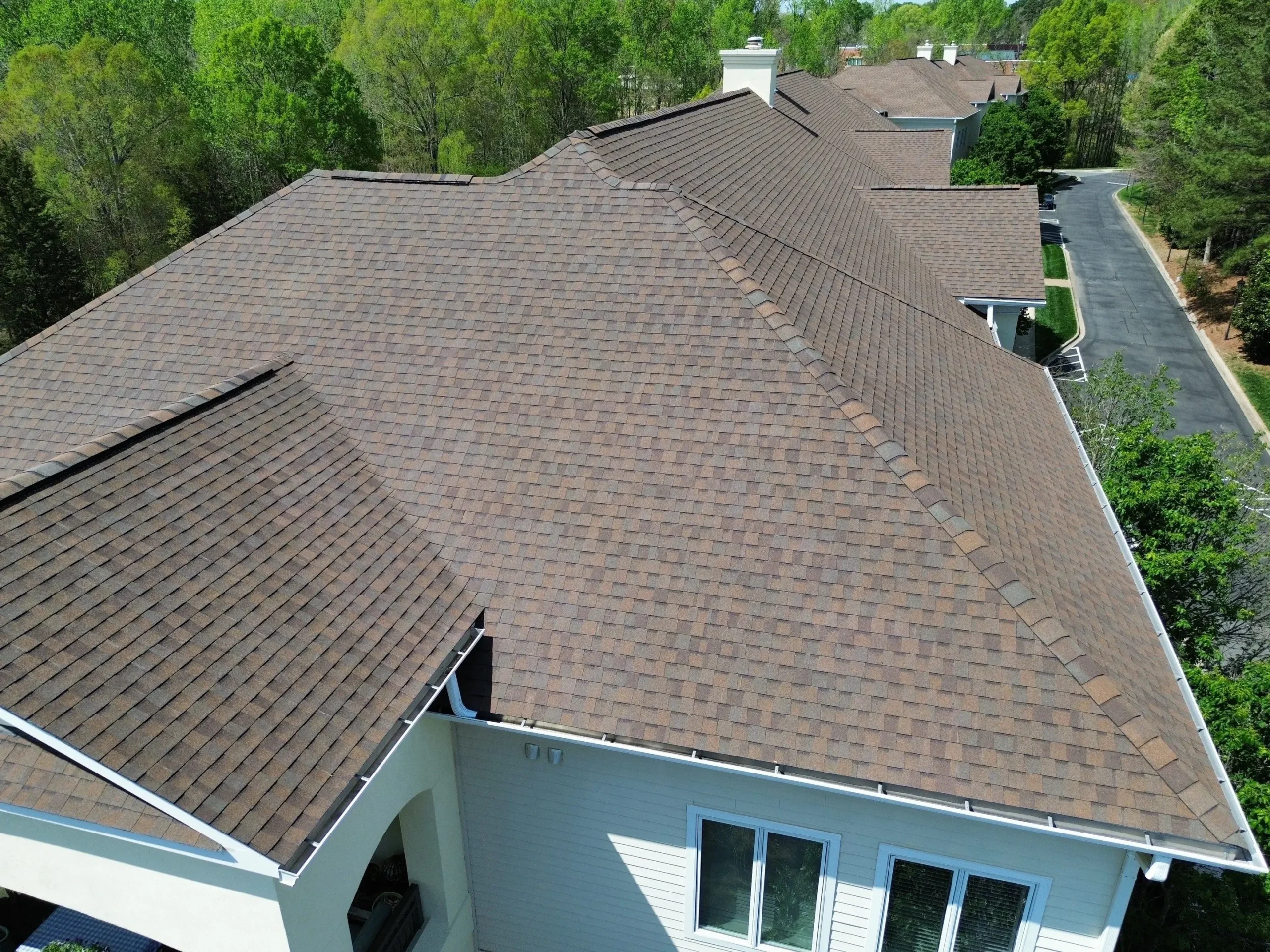 Aerial view of a large building with Brava Cedar Shake brown shingle roofing, surrounded by green trees and a paved road; white exterior walls are visible.