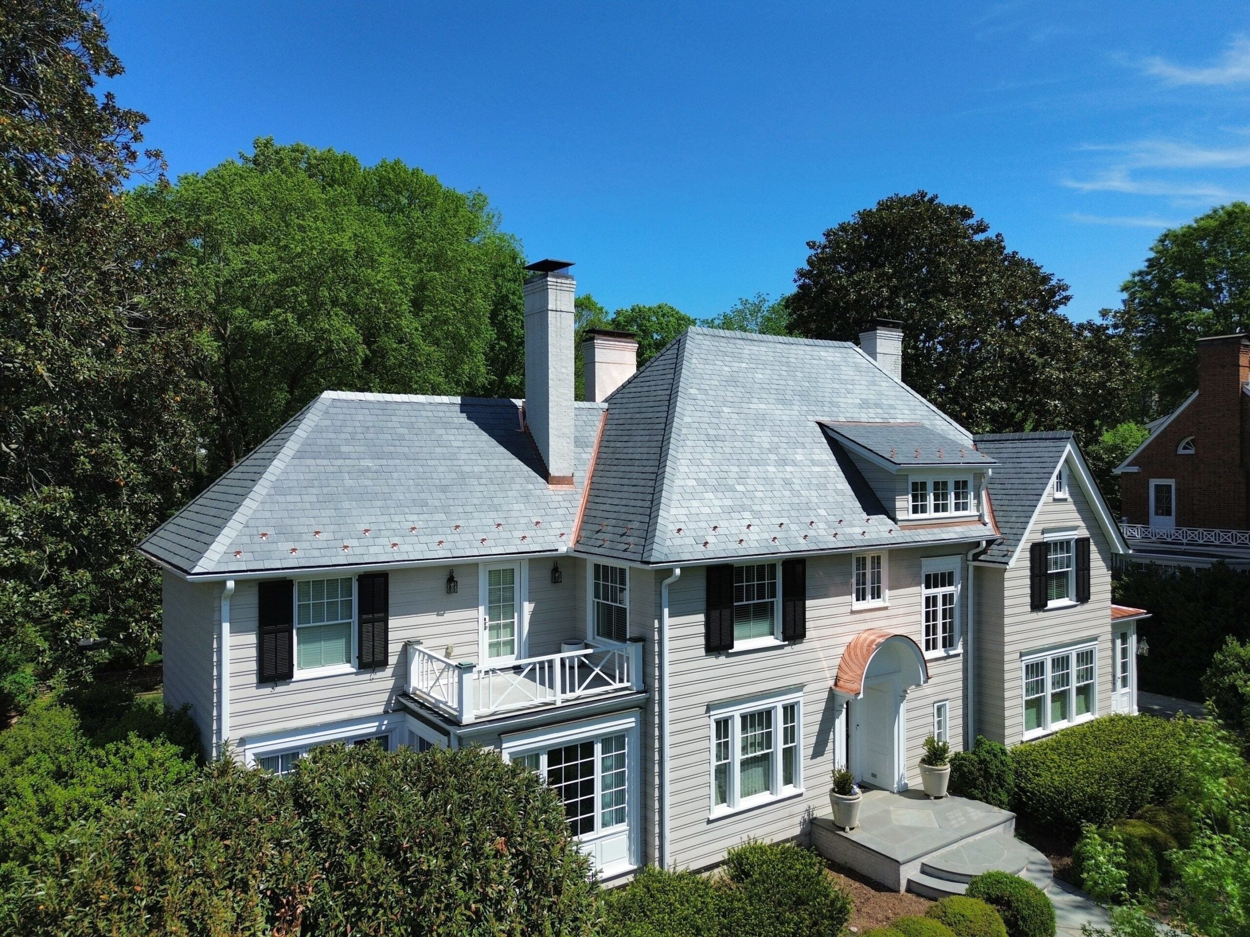 Large two-story white house with dark shutters, a grey roof recently completed by a roof replacement Mooresville NC specialist, multiple chimneys, and a small balcony, surrounded by green trees and bushes under a clear blue sky.