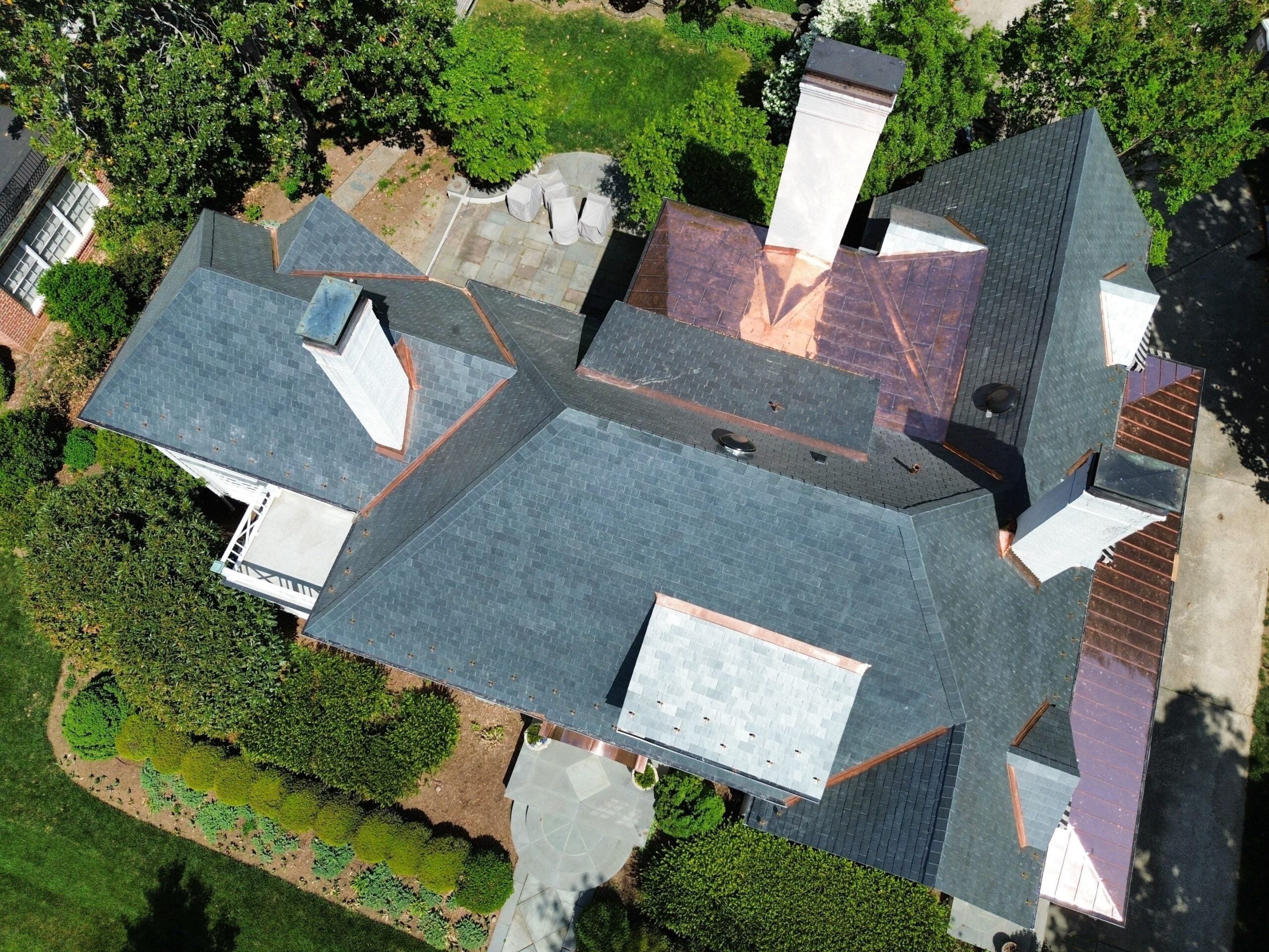 Aerial view of a large house with a dark gray roof, multiple chimneys, and a patio area surrounded by green trees and bushes—an ideal showcase for quality roof replacement. Pathways and landscaped gardens are visible around the property, highlighting the beautiful setting in Mooresville NC.