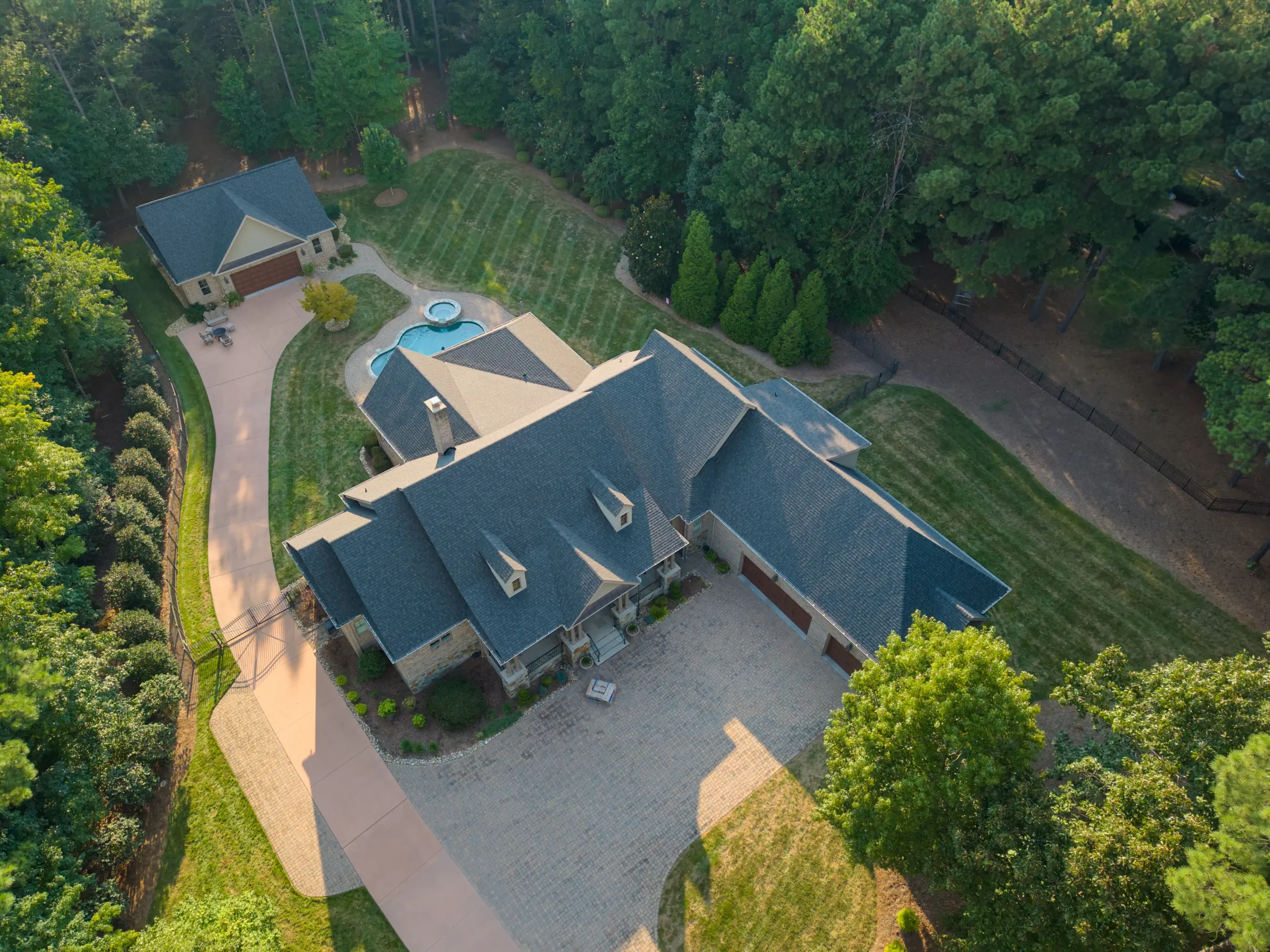 Aerial view of a large house with a gray roof, spacious driveway, separate garage, swimming pool, and well-maintained lawn, all surrounded by dense green trees—an ideal property for those considering roof replacement in Mooresville NC.
