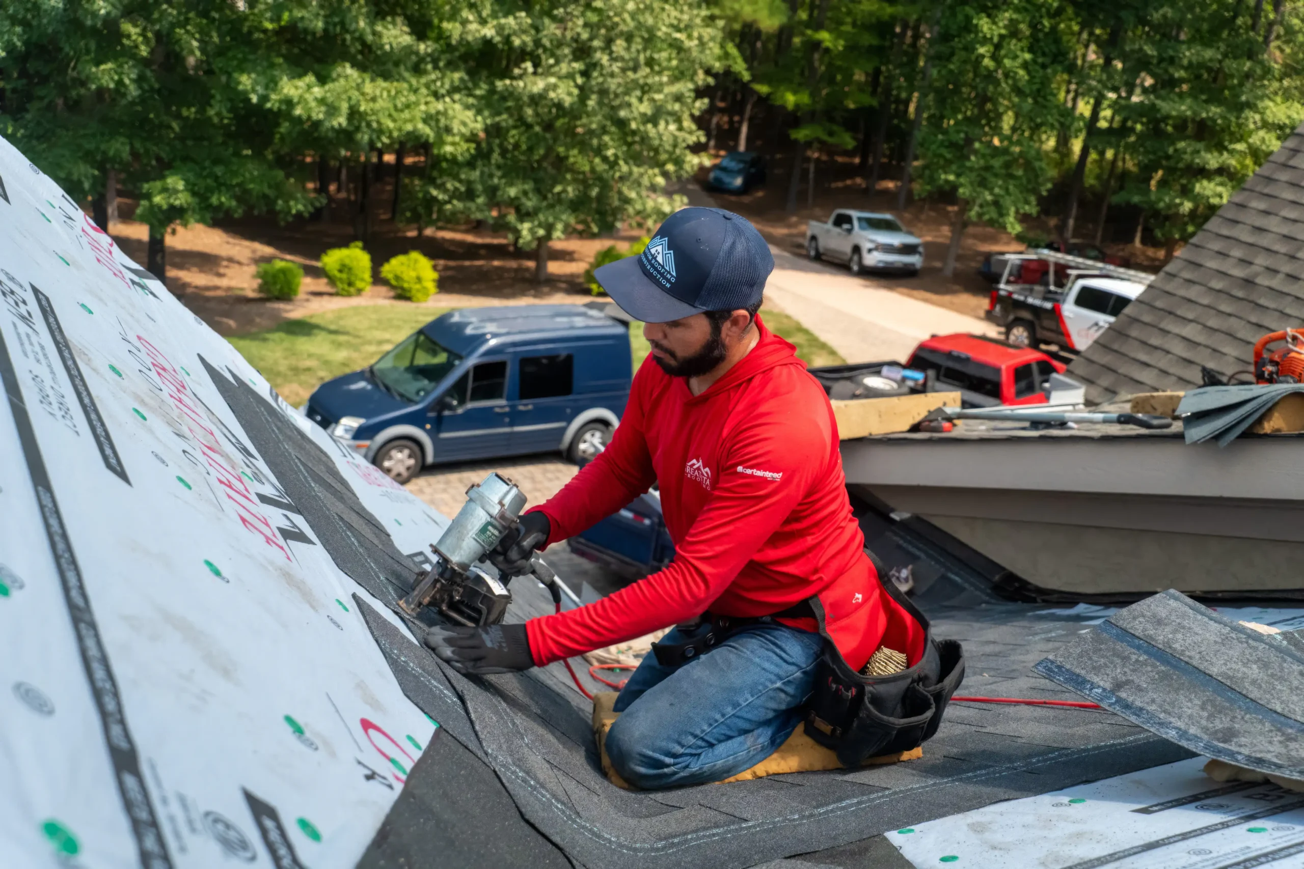 A worker in a red shirt and cap uses a nail gun to install roofing material on a house during a roof replacement in Mooresville, NC, surrounded by trees and parked vehicles.