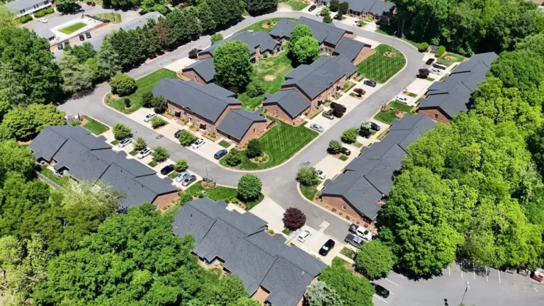 Aerial view of a suburban neighborhood with several houses, possibly managed by an HOA roof replacement Rock Hill SC, arranged around a central grassy area, surrounded by roads, trees, and parked cars.