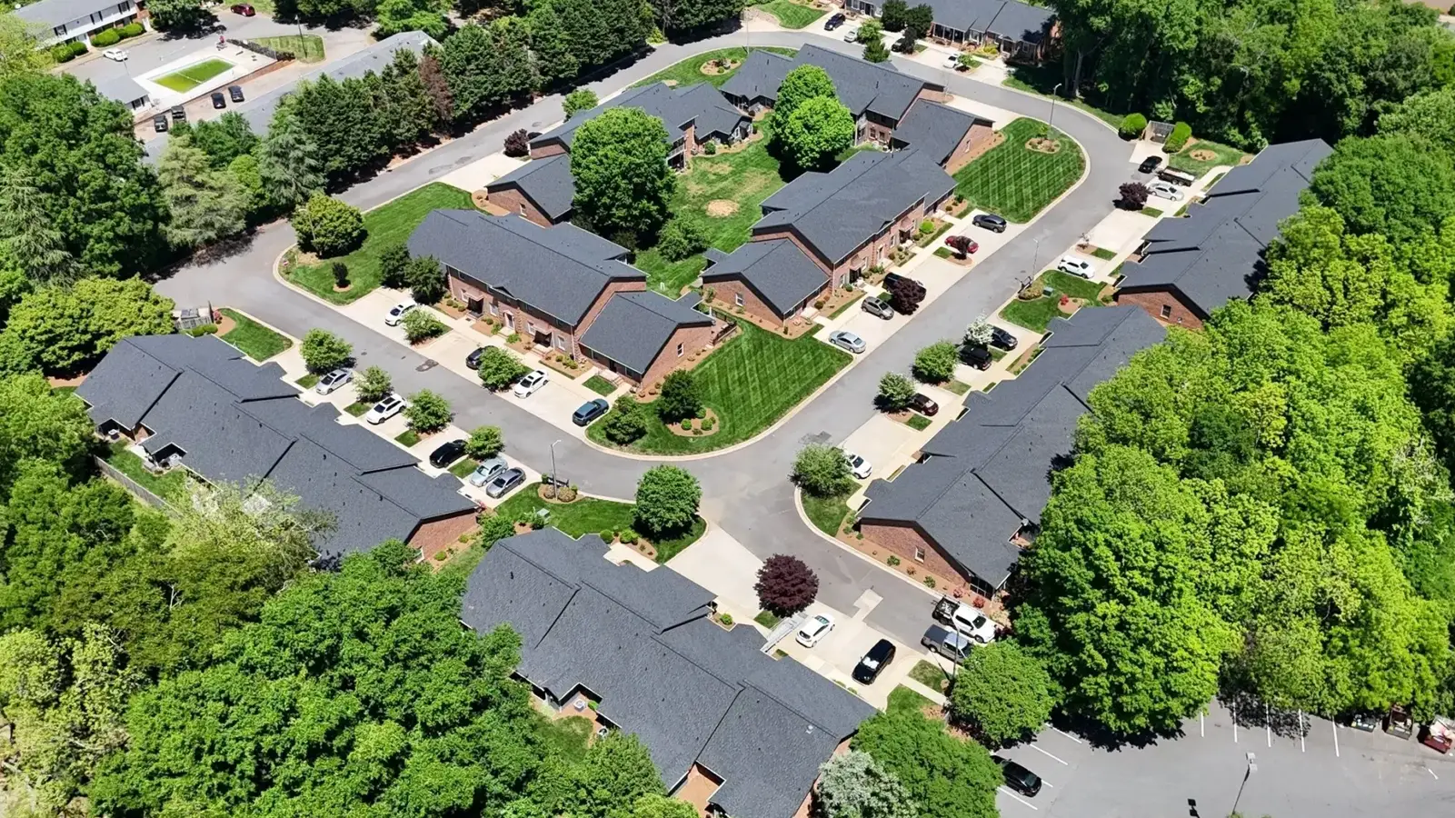 Aerial view of a suburban neighborhood with several houses, possibly managed by an HOA roof replacement Rock Hill SC, arranged around a central grassy area, surrounded by roads, trees, and parked cars.