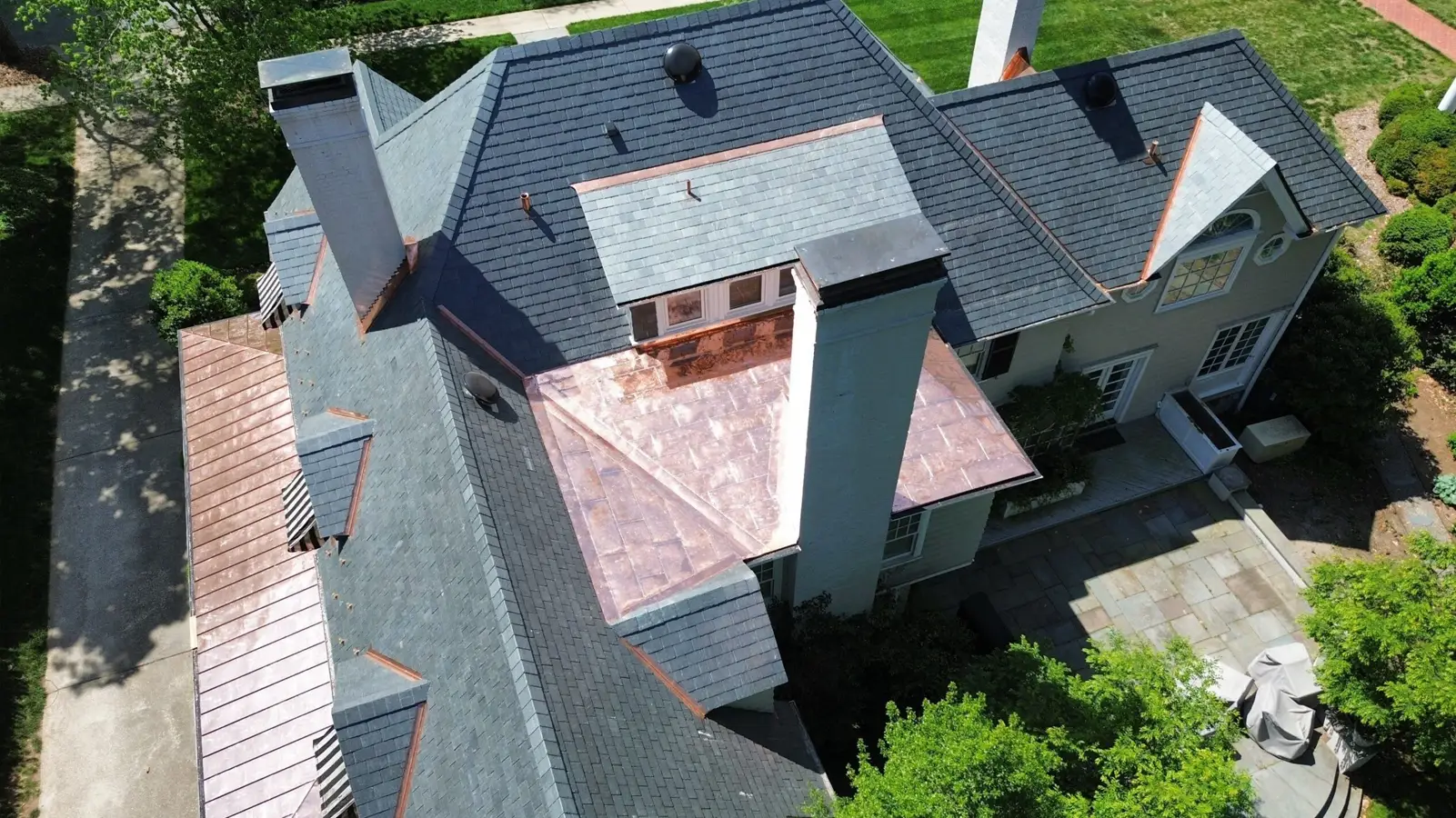 Aerial view of a house with gray shingle roofs, copper flashing, multiple chimneys, and a stone patio surrounded by greenery after a recent roof replacement Mooresville NC.