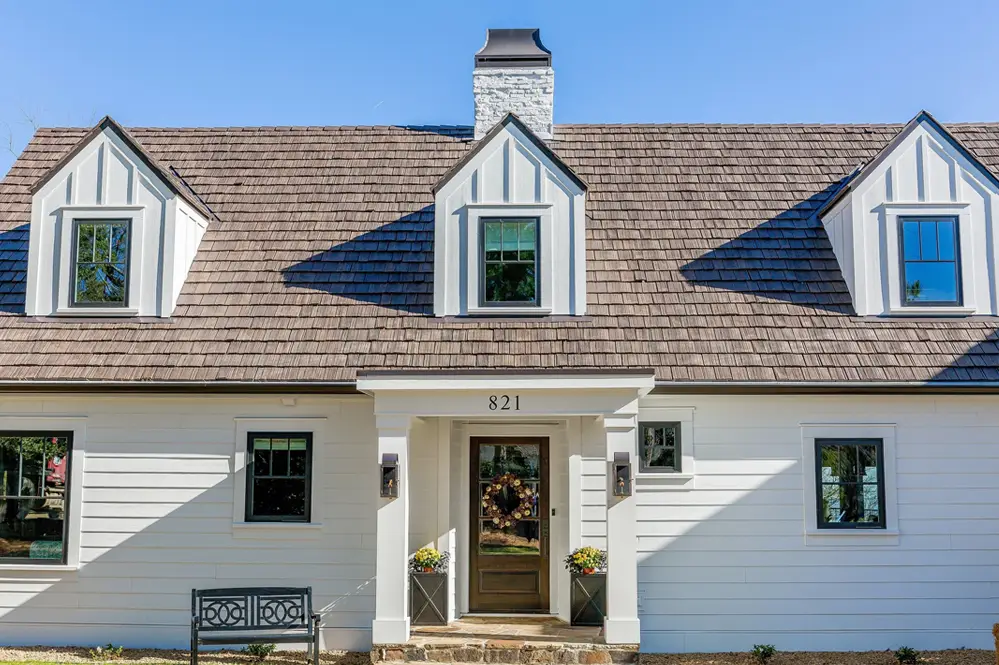 White house with a Brava Composite Roofing brown shingle roof, three dormer windows, central chimney, and a front door flanked by windows and flower boxes. The house number 821 is above the door.