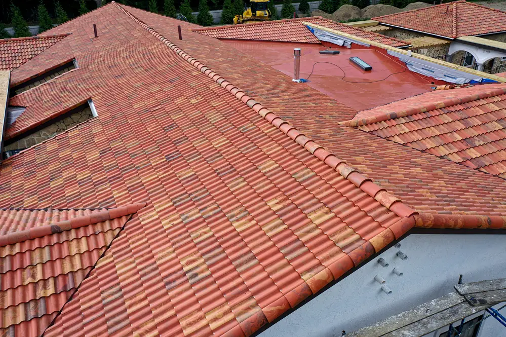 A large rooftop with reddish-brown Brava Composite Roofing tiles, featuring multiple slopes and ridges. One section has a flat red surface with construction materials and tools scattered on it.