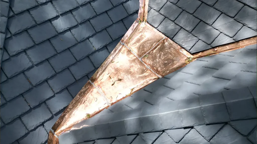 A copper roof valley flashing set between dark gray composite slate roof tiles.