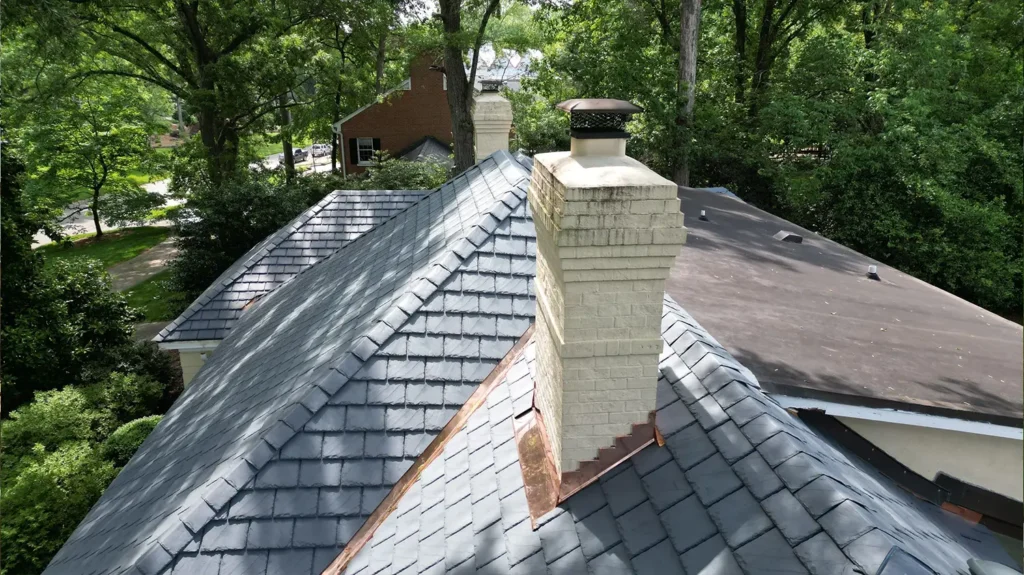 A light-colored brick chimney rises from a composite slate roof, surrounded by mature green trees.