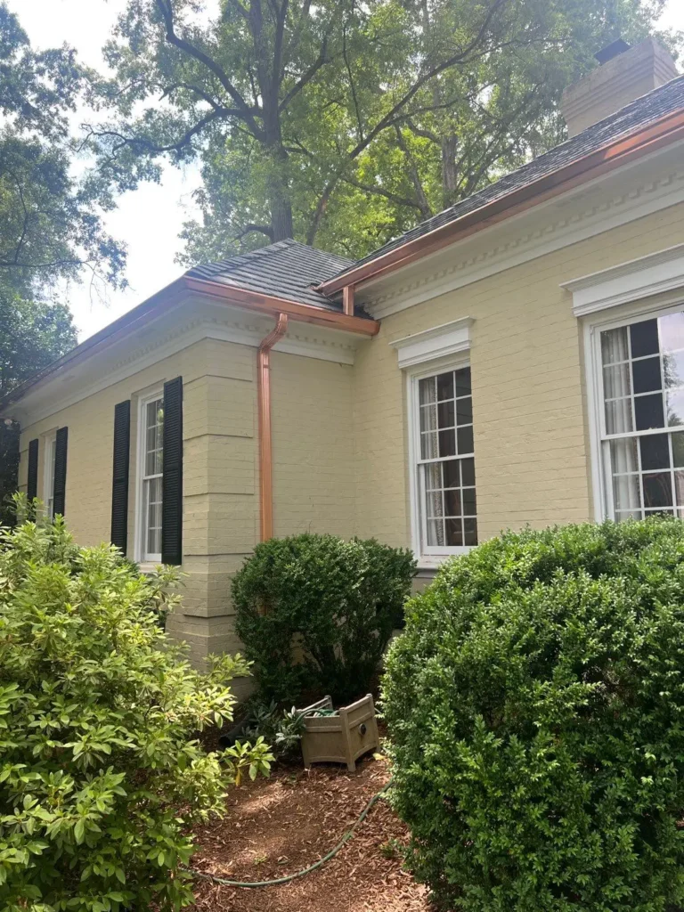 A yellow brick house with new copper gutters and downspouts, surrounded by shrubs and trees, following a recent composite slate roof replacement in Charlotte NC.