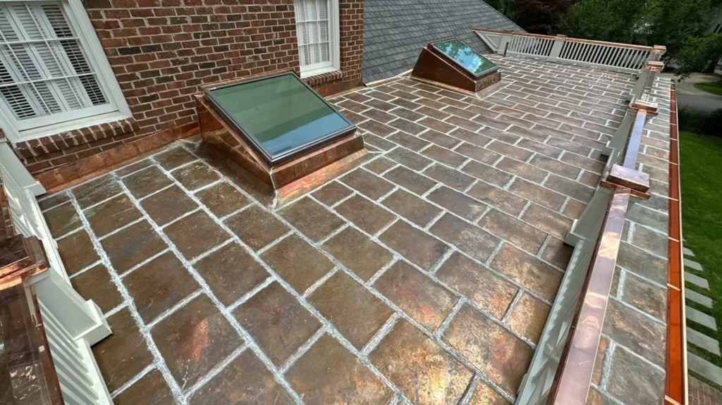 A flat rooftop covered in rectangular copper tiles with two skylights, bordered by a white railing and a brick wall.