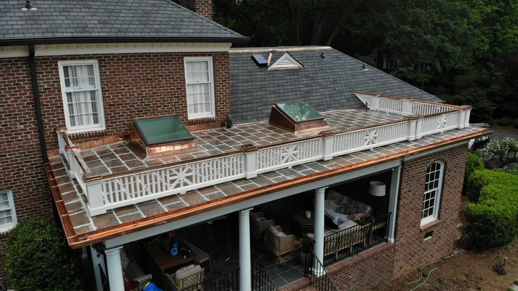 Aerial view of a brick house with a flat stone-tiled roof terrace enclosed by a white wooden railing and copper flashing, featuring two skylights.