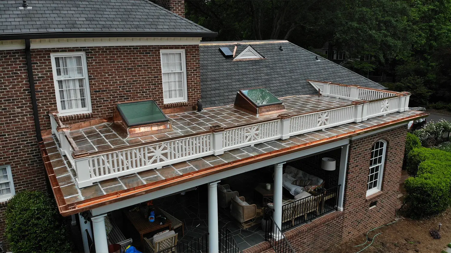 Aerial view of a brick house with a flat stone-tiled roof terrace enclosed by a white wooden railing and copper flashing, featuring two skylights.