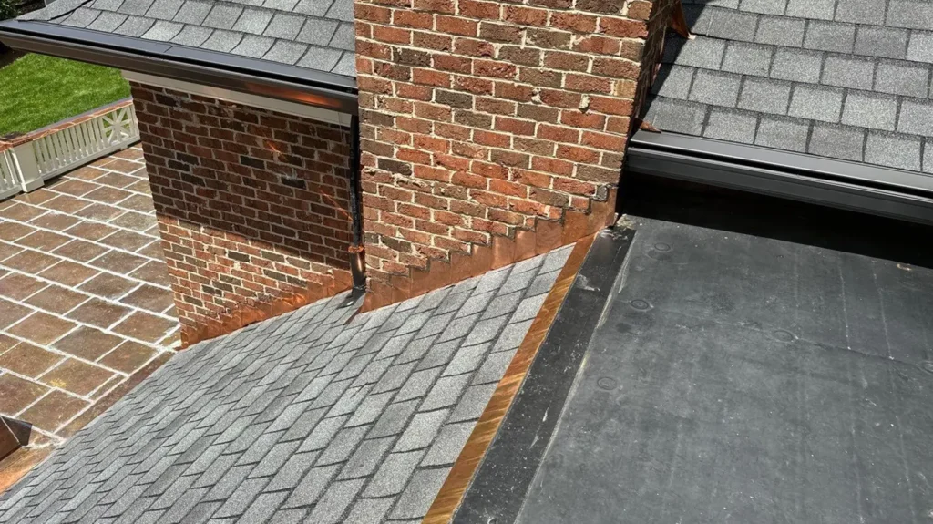 A brick chimney with custom copper flashing on a roof featuring both asphalt shingles and flat roofing material, installed by Charlotte roofing contractors.