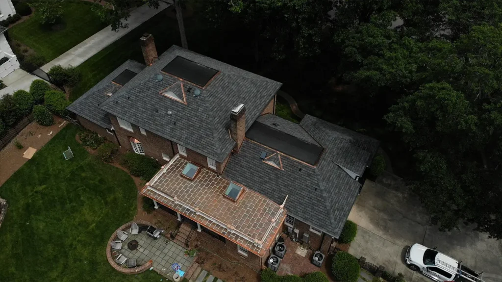 Aerial view of a brick house featuring GAF Slateline roof replacement, skylights, a flat-roofed terrace, and a circular stone patio with outdoor seating.